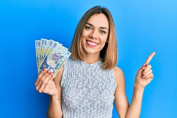Young caucasian blonde woman holding 100 romanian leu banknotes smiling happy pointing with hand...