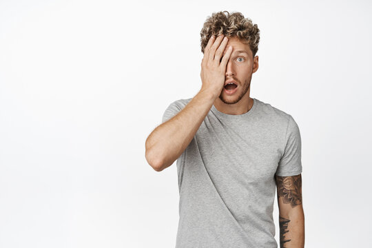Shocked blond man facepalm, staring in awe at camera, drop jaw and looking in disbelief at advertisement, standing in gray t-shirt over white background