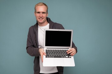 Photo of handsome blond man holding computer laptop looking at camera isolated over blue wall background wearing white t-shirt and grey sweater