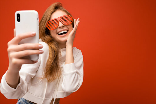 Closeup portrait of attractive positive smiling young blonde woman wearing stylish white hoodie and funny colorful glasses standing isolated over colourful background holding and using mobile phone