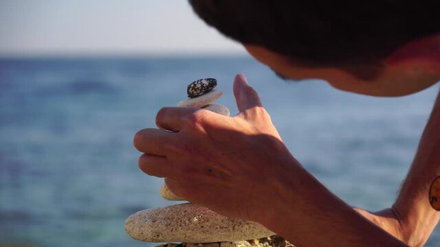 Man bilds stones pyramid on the seashore on a sunny day on the blue sea background. Happy holidays. Pebble beach, calm sea, travel destination. Concept of happy vacation on the sea, meditation, spa