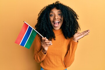 African american woman with afro hair holding gambia flag celebrating achievement with happy smile and winner expression with raised hand - obrazy, fototapety, plakaty