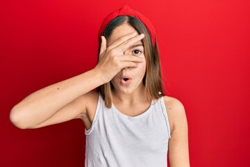 Beautiful brunette little girl wearing casual white t shirt peeking in shock covering face and eyes with hand, looking through fingers afraid