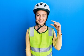 Beautiful brunette little girl wearing bike helmet and reflective vest smiling and confident gesturing with hand doing small size sign with fingers looking and the camera. measure concept.