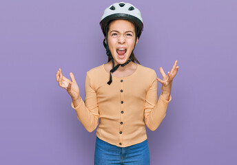 Beautiful brunette little girl wearing bike helmet crazy and mad shouting and yelling with aggressive expression and arms raised. frustration concept.