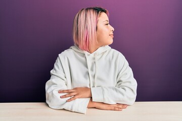 Hispanic woman with pink hair wearing casual sweatshirt sitting on the table looking to side, relax profile pose with natural face with confident smile.
