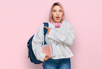 Hispanic woman with pink hair wearing student backpack and headphones surprised pointing with finger to the side, open mouth amazed expression.