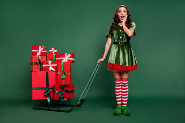 Full length body size view of pretty cheerful amazed girl elf bringing gifts isolated over green...