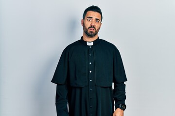 Handsome hispanic man with beard wearing catholic priest robe in shock face, looking skeptical and sarcastic, surprised with open mouth