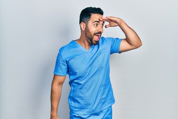 Fototapeta premium Handsome hispanic man with beard wearing blue male nurse uniform very happy and smiling looking far away with hand over head. searching concept.