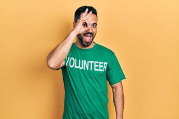 Handsome hispanic man with beard wearing volunteer t shirt smiling happy doing ok sign with hand on eye looking through fingers