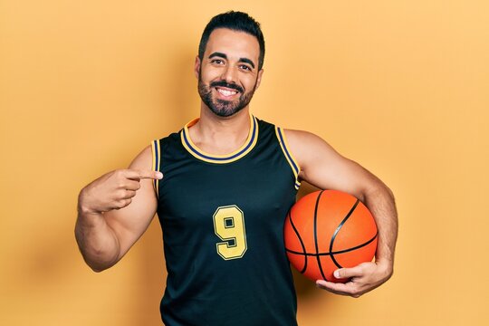 Handsome hispanic man with beard holding basketball ball pointing finger to one self smiling happy and proud