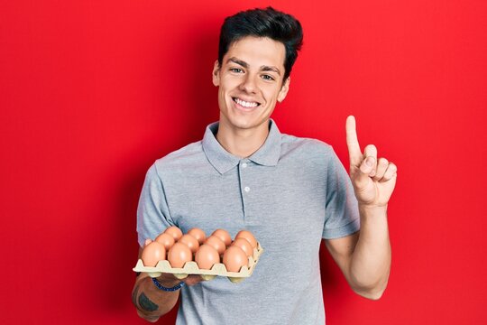 Young hispanic man showing fresh white eggs smiling with an idea or question pointing finger with happy face, number one