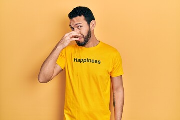 Hispanic man with beard wearing t shirt with happiness word message smelling something stinky and disgusting, intolerable smell, holding breath with fingers on nose. bad smell