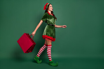 Full length body size view of attractive cheerful girl elf carrying bags gifts isolated over green color background