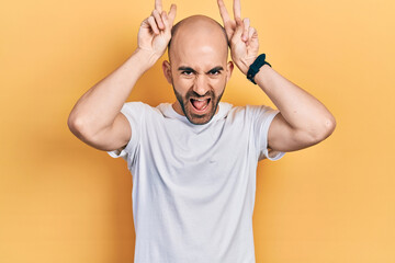 Young bald man wearing casual white t shirt posing funny and crazy with fingers on head as bunny ears, smiling cheerful