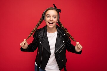 Shot of attractive positive smiling brunette little female teenager with pigtails wearing stylish black leather jacket and white t-shirt isolated over red background wall looking at camera