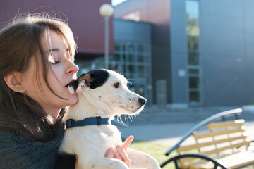 young woman sitting on a bench in the city and holding jack russel terrier . woman kissing dog's ear while he looking aside. happy pet owner resting outdoors in the sunlight.
