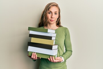 Young blonde woman holding a pile of books relaxed with serious expression on face. simple and natural looking at the camera.