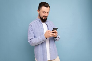 Photo shot of handsome positive good looking young man wearing casual stylish outfit poising isolated on background with empty space holding in hand and using mobile phone messaging sms looking at