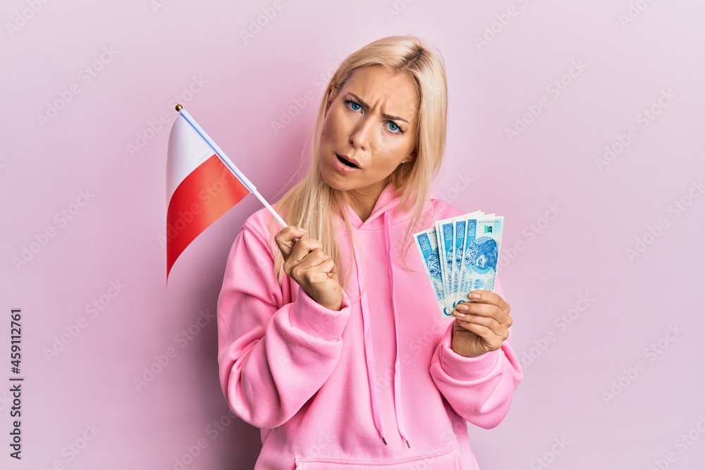 Canvas Prints young blonde woman holding poland flag and zloty banknotes in shock face, looking skeptical and sarc