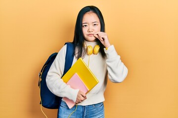 Young chinese girl holding student backpack and books mouth and lips shut as zip with fingers....