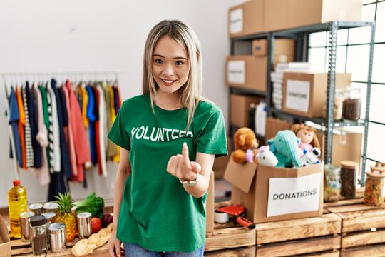 Asian young woman wearing volunteer t shirt at donations stand beckoning come here gesture with hand inviting welcoming happy and smiling