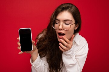 photo of sexy amazed beautiful young brunette woman wearing white shirt and optical glasses isolated over red background holding in hand and showing mobile phone with empty screen for cutout looking