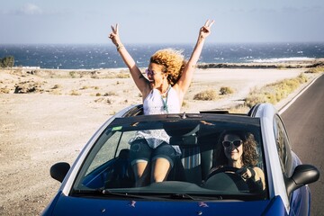 Fototapeta premium Two young women traveling in a convertible car and having fun during journey. Young woman posing with v sign while friend driving car on road. Two female friends having fun on car road trip
