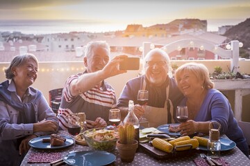 Cheerful two senior couples dining at terrace party and taking selfie using smartphone. Elderly people having fun while dining at rooftop. Old family photographing using mobile phone in celebration