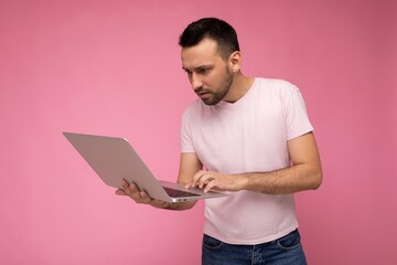 Handsome brunet man holding laptop computer and typing on the keyboard looking at netbook monitor in t-shirt on isolated pink background