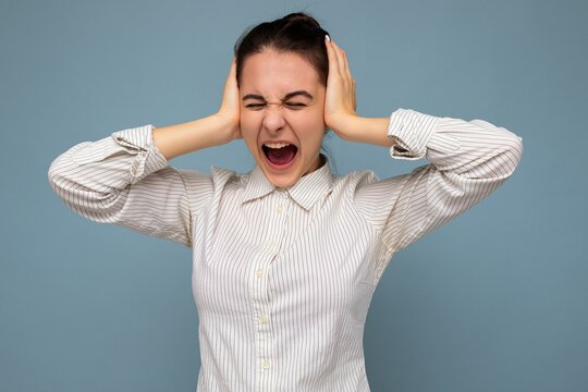 Portrait Of Emotional Young Beautiful Brunette Woman With Sincere Emotions Wearing White Shirt Isolated Over Blue Background With Copy Space And Covering Ears With Hands And Shouting