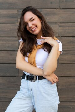 Vertical ptoto of young beautiful positive happy brunette woman standing against brown wall in the street and wearing stylish outfit