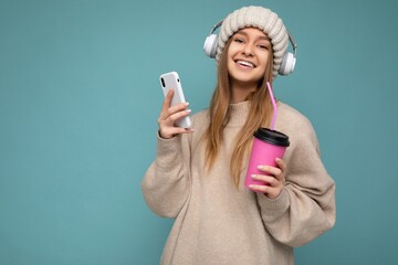 Attractive smiling young blonde woman wearing beige sweater and beige hat white headphones isolated over blue background holding in hand and using mobile phone drinking beverage and listening to music