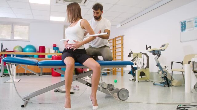 Handheld shot of a young Caucasian woman during rehab treatment, and male physiotherapist helping her to do back and hip exercises with medical bar properly