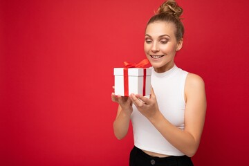 Photo shot of beautiful positive smiling surprised young blonde woman isolated over red background wall wearing white top holding gift box and looking at present