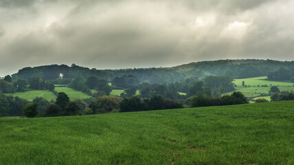 Hilly landscape with meadows and trees under a cloudy sky.