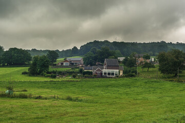 Houses in a hilly landscape with trees and meadows under a cloudy sky.