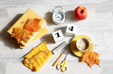 Calendar for October 14 : the name of the month in English, cubes with the number 14, books, an alarm clock, maple leaves, a knitted scarf, a cup of coffee, eyeglasses on a gray background