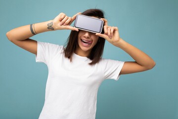Photo of sexy pretty positive young brunette woman good looking wearing white t-shirt standing isolated on blue background with copy space holding smartphone showing phone in hand with empty screen