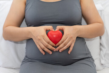 Pregnant woman holding red heart touching her belly. Loving mom waiting of a baby. Maternity, parenting, prepare and expect concept. 