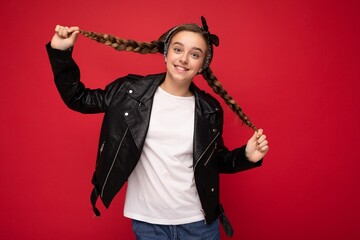 Shot of pretty positive smiling brunette little female teenager with pigtails wearing stylish black leather jacket and white t-shirt for mockup standing isolated over red background wall looking at
