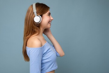 Side-profile photo of beautiful positive smiling young blonde woman wearing blue crop top isolated over blue background wall wearing white wireless bluetooth headphones listening to cool music and