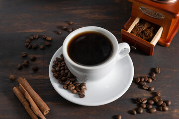 A white cup with black coffee on a dark wooden table, coffee beans on a saucer. Cinnamon sticks and a coffee grinder in the background