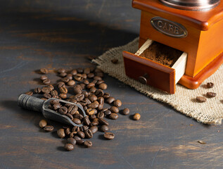 Coffee beans lie in a wooden spoon and are scattered on a dark table. Coffee grinder in the background