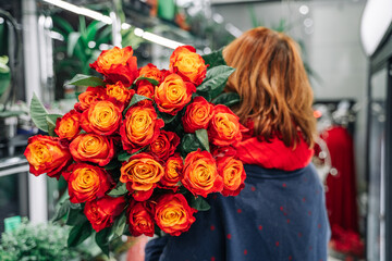 Red-haired woman florist holding a luxurious bouquet with fresh red-orange roses. Flower show in the store.
