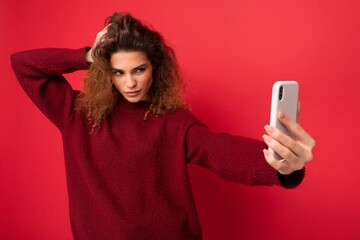 Sexy Attractive young blogger woman with curly hair wearing dark red sweater isolated on red...