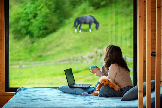 Woman sitting on the bed and looks outside the window seeing mountain. Concept of the workplace at mountain chalet, working remotely.
