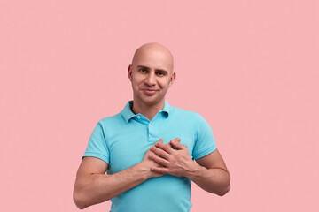 Believe me. Sincere bald homosexual man with bristle falls in love, presses hands to heart, keeps palms on chest, smiles, tilts head, gay friendly, wears blue polo shirt, stands over pink background.