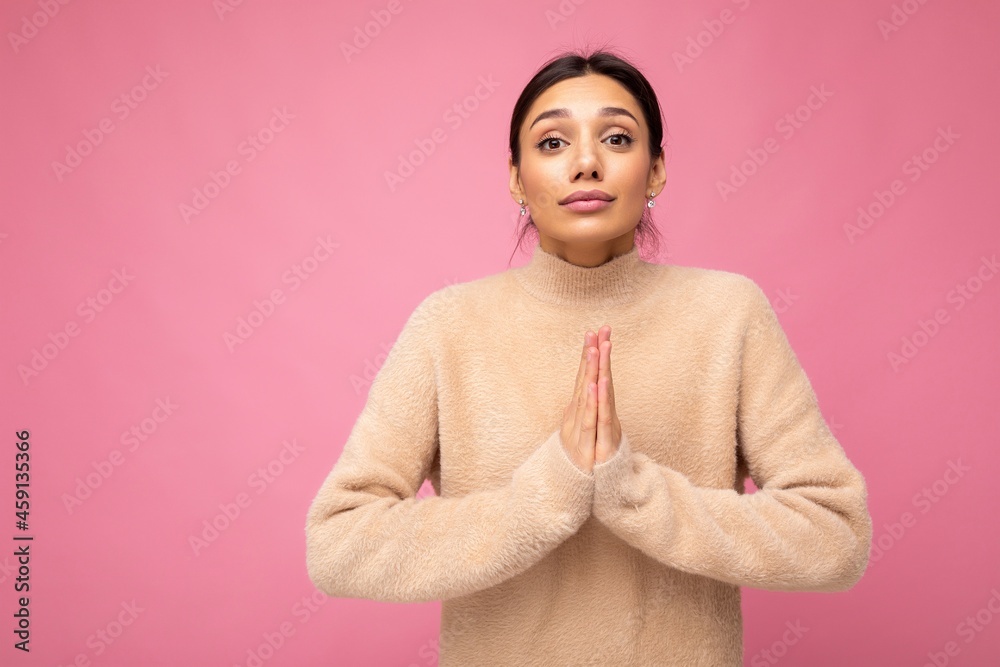 Wall mural Portrait of young serious beautiful brunette woman with sincere emotions wearing casual beige jersey isolated over pink background with free space and keeping hands together asking for forgiveness